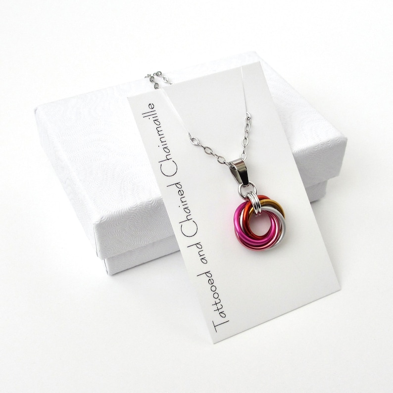 May include: A silver chain necklace with a pendant made of interlocking rings in pink, gold, and silver. The necklace is displayed on a white card with the text "Tattooed and Chained Chainmaille" and a white gift box.