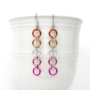 May include: A pair of long dangle earrings with a gradient of colors. The earrings are made of metal rings in shades of orange, gold, silver, and pink.