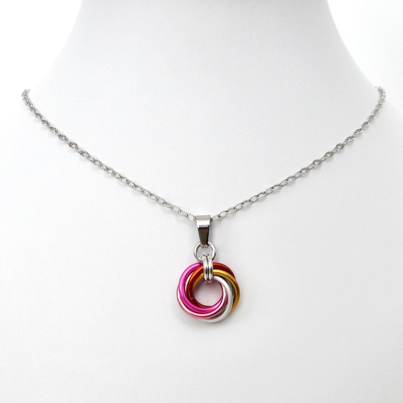 May include: A silver-toned necklace with a pendant featuring a colorful interlocking ring design. The pendant incorporates shades of pink, gold, and silver, suspended from a delicate chain. The necklace is displayed on a white mannequin.