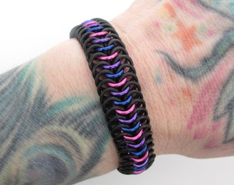May include: A black, blue, and pink chainmail bracelet. The bracelet is made of small metal rings that are linked together in a pattern.