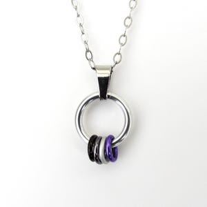 May include: A silver-toned necklace with a large circular pendant. Three smaller rings in black, gray, and purple are attached to the bottom of the larger ring. The necklace chain is made of small, linked circles.