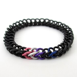 K&ouml;nnte beinhalten: Ein schwarzes Kettenhemdarmband mit gewebtem Design. Das Armband hat einen kleinen Anh&auml;nger mit rosa, wei&szlig;en und blauen Ringen.