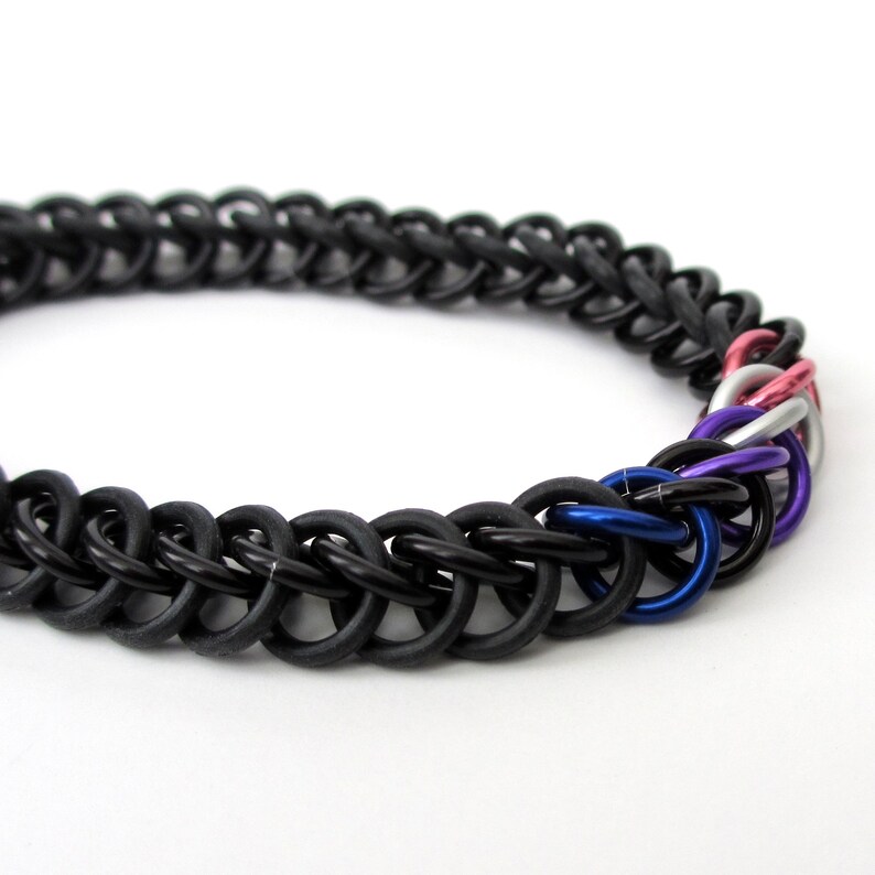 K&ouml;nnte beinhalten: Ein schwarzes Kettenhemdarmband mit einem gewebten Design und einem mehrfarbigen Akzent. Der Akzent besteht aus Ringen in Pink, Wei&szlig;, Blau und Lila.