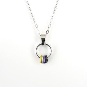 May include: A silver-colored necklace with a large circular pendant. Three smaller rings in yellow, black, and purple are attached to the bottom of the pendant. The necklace chain is also silver and hangs against a white background.