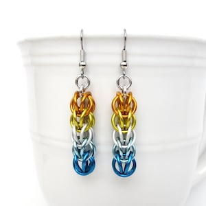 May include: A pair of dangle earrings with a graduated color pattern. The earrings are made of metal and have a silver hook. The top of the earrings are orange, then yellow, then silver, then blue.