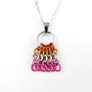 May include: A silver chain necklace with a pendant featuring a series of colorful metal rings. The rings are arranged in rows of orange, gold, silver, and pink. The pendant is attached to a larger silver ring.