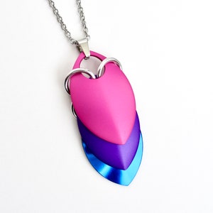 Peut inclure: Un collier avec un pendentif en forme de cœur superposé, dans les tons rose, violet et bleu. Le pendentif est en métal et est sur une chaîne argentée.
