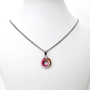 May include: A silver chain necklace with a pendant featuring a colorful, interlocking ring design. The pendant incorporates pink, gold, and silver-colored metal rings. The necklace is displayed on a white mannequin.