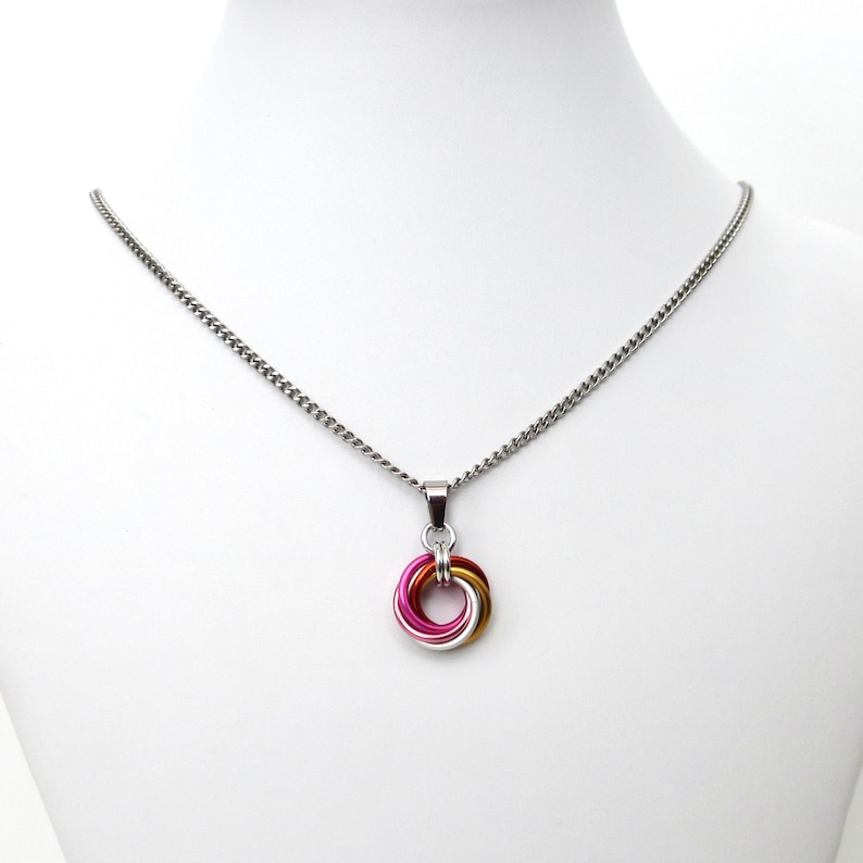 May include: A silver chain necklace with a pendant featuring a colorful, interlocking ring design. The pendant incorporates pink, gold, and silver-colored metal rings. The necklace is displayed on a white mannequin.