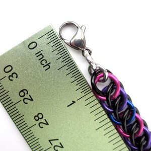 May include: A close-up of a black, purple, and pink chainmail bracelet with a silver lobster clasp. The bracelet is measured against a ruler, showing it is just over 1 inch long.