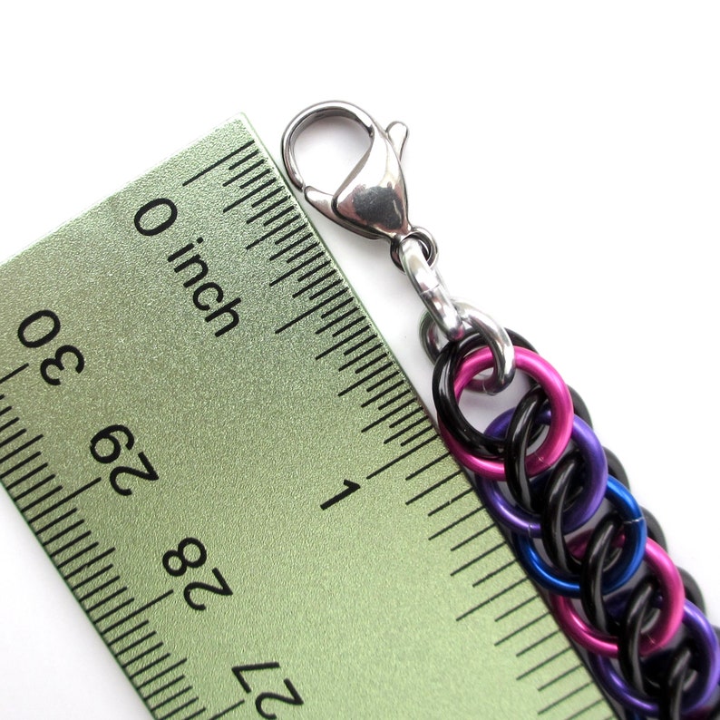 May include: A close-up of a black, purple, and pink chainmail bracelet with a silver lobster clasp. The bracelet is measured against a ruler, showing it is just over 1 inch long.