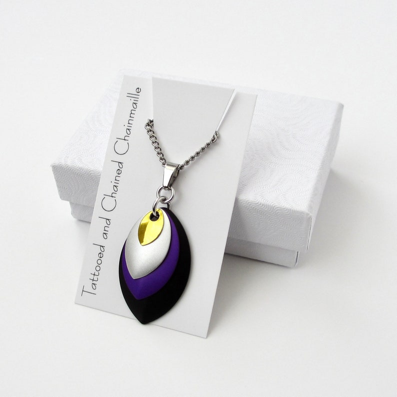 K&ouml;nnte beinhalten: Eine silberne Kettenkette mit einem mehrschichtigen Anh&auml;nger aus schwarzem, violettem, silbernem und goldenem Metall. Der Anh&auml;nger ist tropfenf&ouml;rmig und befindet sich auf einer wei&szlig;en Karte mit dem Text "Tattooed and Chained Chainmaille".