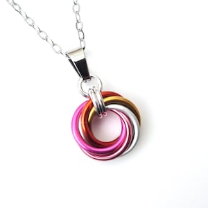 May include: A silver chain necklace with a pendant featuring interlocking rings in shades of pink, red, gold, and silver. The pendant is made of metal and has a bright, reflective finish. The necklace is displayed against a white background.