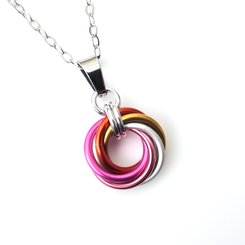 May include: A silver chain necklace with a pendant featuring interlocking rings in shades of pink, red, gold, and silver. The pendant is made of metal and has a bright, reflective finish. The necklace is displayed against a white background.