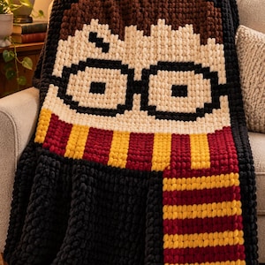 May include: A chunky knit blanket featuring a pixelated character design with brown hair, round black glasses, and a lightning bolt scar. The character wears a scarf with horizontal stripes in deep red and yellow, set against a black background.