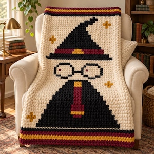 May include: A chunky knit throw blanket in cream, featuring a pixelated graphic of a figure wearing a black pointed hat with a maroon band, round glasses, and a maroon and gold tie, accented by small gold cross-shaped stars and striped borders.
