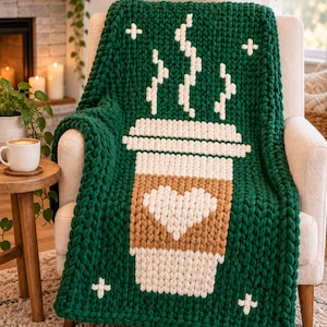 Op de afbeelding: Een grof gebreide plaid in donkergroen met een grote, centrale afbeelding van een koffiebeker met een beige hoes, een wit hart en witte stoom die erboven uitstijgt. De deken is voorzien van kleine witte kruisvormige accenten in de hoeken.