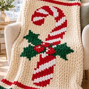 May include: A chunky knit cream-coloured throw blanket featuring a large red and white striped candy cane design in the centre, accented with green holly leaves and red berries. The blanket has thick green and red striped borders.