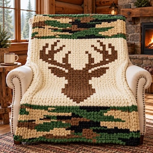 May include: A chunky knit throw blanket featuring a large brown deer head silhouette centred on a cream background, framed by top and bottom bands of camouflage pattern in shades of dark green, brown, black, and tan yarn textures.