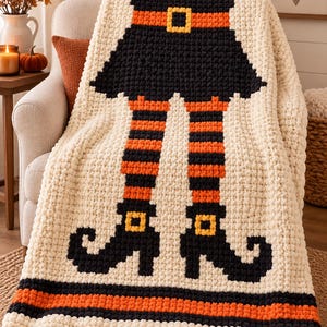 May include: A cream-colored crochet blanket featuring a central graphic of a witch's legs in orange and black striped stockings, wearing black pointed shoes with yellow buckles, and a black skirt with an orange belt and yellow buckle.