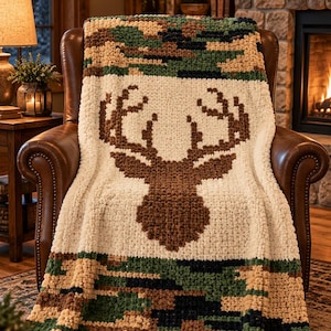 May include: A chunky knit blanket featuring a central brown deer head silhouette on a cream background, bordered by top and bottom sections of green, brown, and black camouflage pattern. The text Loops & Co. appears in the top left corner.