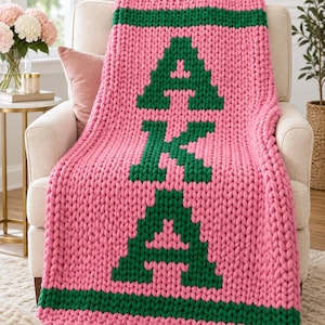 Peut inclure: Un plaid en grosse maille rose vif avec des bandes horizontales vert foncé sur les bords supérieur et inférieur. Le centre du plaid présente les grandes lettres A, K et A en style bloc, tricotées avec un fil de couleur vert foncé.
