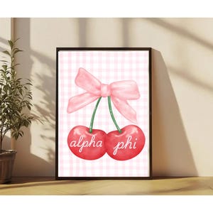 Puede incluir: Impresión artística enmarcada con una ilustración de acuarela de dos cerezas rojas con tallos verdes y un lazo rosa. El texto "alpha phi" está escrito en las cerezas. El fondo es un patrón de cuadros rosa y blanco.