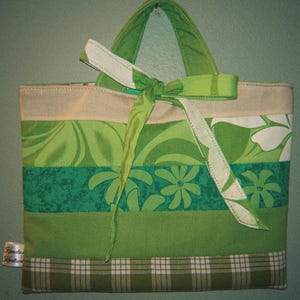 Puede incluir: Bolso tote hecho a mano con un diseño patchwork en varios tonos de verde y beige. El bolso presenta un estampado floral, una base a cuadros y un lazo verde. Las asas son verdes y tiene una pequeña etiqueta con texto adjunta. Altura aproximada: 25 cm.