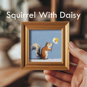 K&ouml;nnte beinhalten: Gerahmtes Gem&auml;lde mit dem Titel "Squirrel With Daisy". Das Kunstwerk zeigt ein braunes Eichh&ouml;rnchen, das eine G&auml;nsebl&uuml;mchen mit wei&szlig;en und gelben Bl&uuml;tenbl&auml;ttern vor einem hellblauen Hintergrund h&auml;lt. Der Rahmen ist goldfarben und quadratisch.