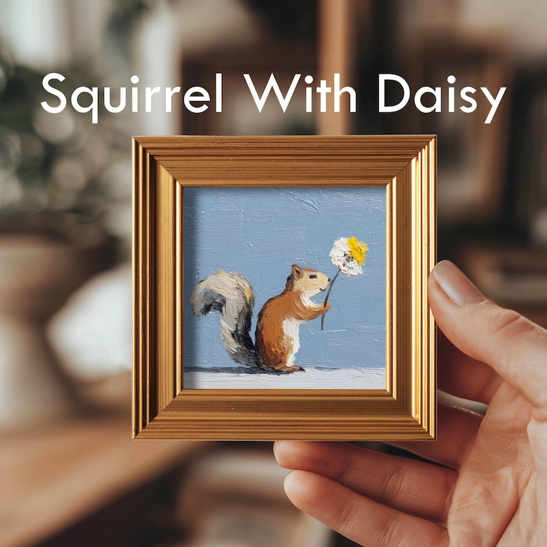 K&ouml;nnte beinhalten: Gerahmtes Gem&auml;lde mit dem Titel "Squirrel With Daisy". Das Kunstwerk zeigt ein braunes Eichh&ouml;rnchen, das eine G&auml;nsebl&uuml;mchen mit wei&szlig;en und gelben Bl&uuml;tenbl&auml;ttern vor einem hellblauen Hintergrund h&auml;lt. Der Rahmen ist goldfarben und quadratisch.