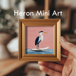 K&ouml;nnte beinhalten: Ein gerahmtes Mini-Kunstwerk mit dem Text "Heron Mini Art". Das Kunstwerk zeigt einen Reiher, der auf einem Surfbrett im Meer steht. Der Hintergrund ist ein rosa Himmel und blaues Wasser. Der Rahmen ist goldfarben.