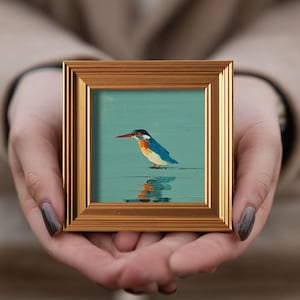 May include: A small, square framed painting of a kingfisher bird. The bird has blue, orange, and white feathers, a long red beak, and is standing in water. The frame is gold-colored.