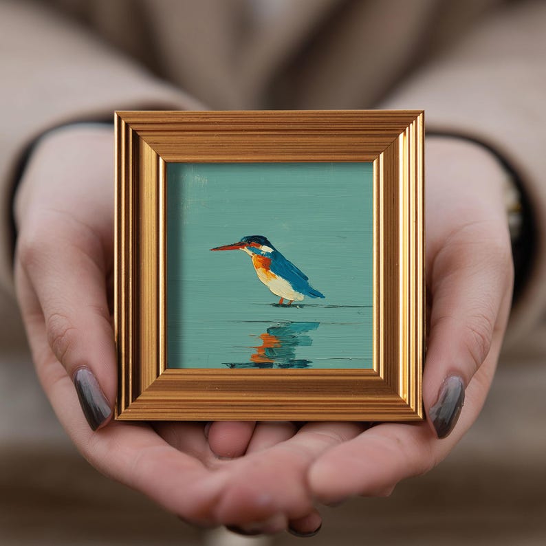 May include: A small, square framed painting of a kingfisher bird. The bird has blue, orange, and white feathers, a long red beak, and is standing in water. The frame is gold-colored.
