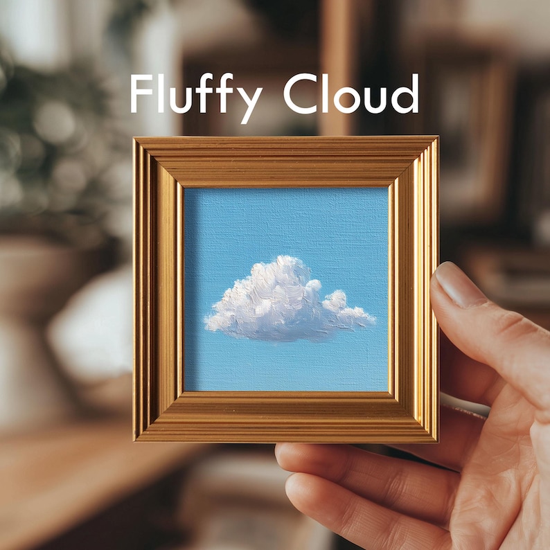May include: A small square painting of a fluffy white cloud against a light blue sky, framed in a gold-colored wooden frame. The text "Fluffy Cloud" is displayed above the painting. The artwork is held by a person's hand.