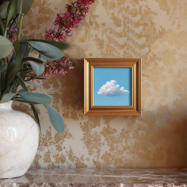 May include: A small, square painting of a fluffy white cloud against a light blue sky, set in a gold frame. The artwork is displayed on a wall with a gold floral pattern, next to a white vase with greenery and pink flowers.