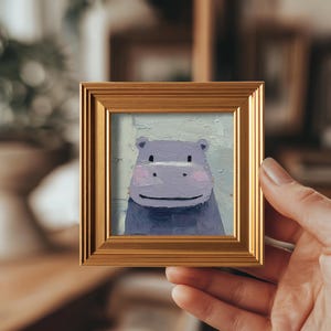 May include: A small, square painting of a smiling hippo in shades of purple and blue, framed in a gold-colored, ornate frame. The hippo has pink cheeks and a happy expression. The artwork is held by a hand.