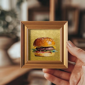 以下が含まれることがあります： ハンバーガーのミニチュア額入り絵画。ハンバーガーは、ゴマのバンズ、ビーフパティ、チーズ、レタス、トマトです。絵画は金色の背景にあり、茶色の木製フレームに額装されています。絵画は手で持たれています。