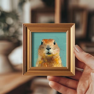 Puede incluir: Pequeña pintura cuadrada de un perro de la pradera en un marco dorado. La obra de arte presenta la cara del perro de la pradera sobre un fondo azul claro. El marco es de color dorado cálido, añadiendo un toque de elegancia a la pieza.