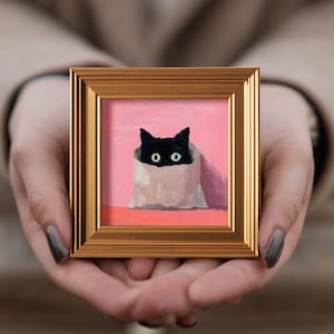 Può includere: Dipinto incorniciato di un gatto nero che sbuca da un sacchetto di carta bianco. Il gatto ha gli occhi verdi ed è ambientato su uno sfondo rosa. La cornice è dorata.