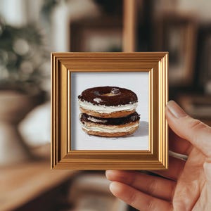 Könnte beinhalten: Ein gerahmtes Miniaturgemälde von zwei übereinander gestapelten, schokoladenüberzogenen Donuts mit weißer Creme-Füllung. Das Gemälde befindet sich in einem goldfarbenen Rahmen und wird von einer Hand gehalten.