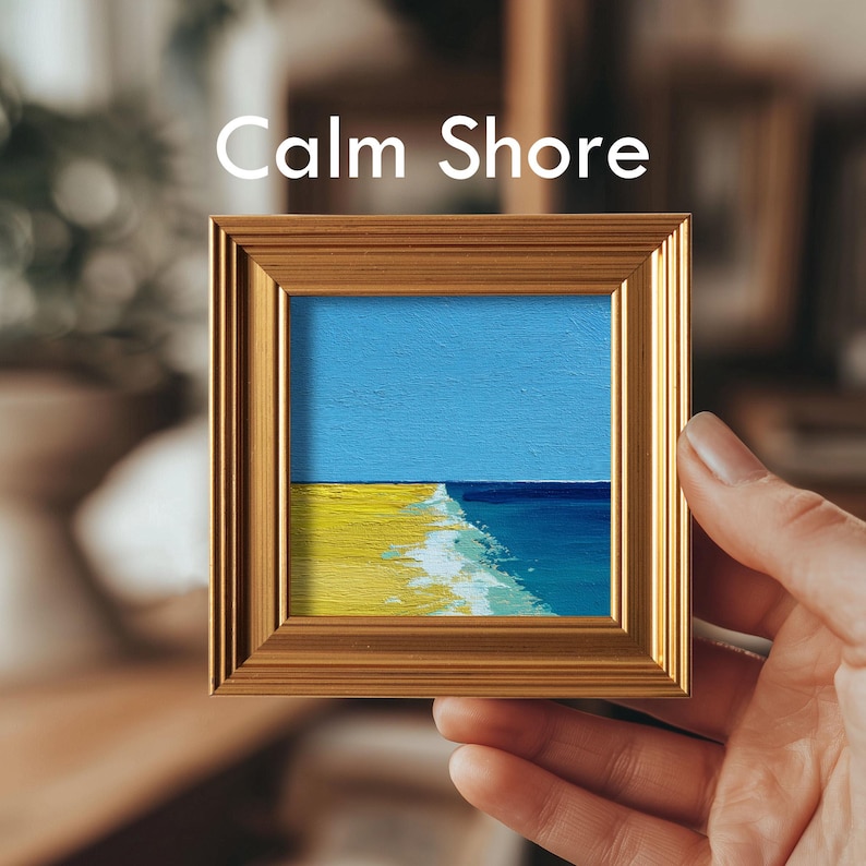 May include: A small framed painting titled "Calm Shore" depicts a beach scene. The artwork features a blue sky, yellow sand, and turquoise water. The painting is in a gold-colored frame and held by a hand.