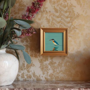 May include: A framed painting of a kingfisher bird with blue, orange, and white plumage, standing in water. The artwork is set against a teal background and framed in a gold square frame. The painting is displayed on a wall with a floral pattern.
