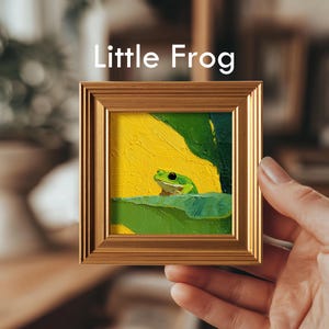 May include: A small framed painting titled "Little Frog" depicts a green frog on a lily pad against a yellow background. The artwork is encased in a gold-colored frame. The painting is held by a hand.