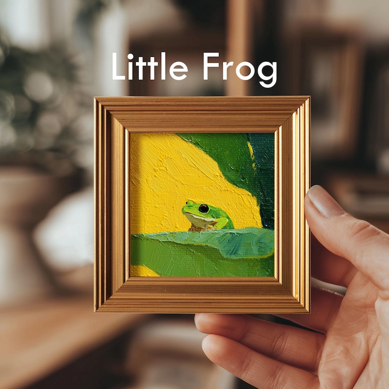 May include: A small framed painting titled "Little Frog" depicts a green frog on a lily pad against a yellow background. The artwork is encased in a gold-colored frame. The painting is held by a hand.