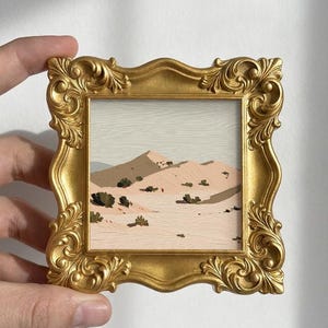May include: A small square painting of a desert landscape with beige sand dunes and dark green shrubs under a pale sky, encased in an ornate, metallic gold-colored frame with intricate scrollwork and decorative floral-like corner carvings.
