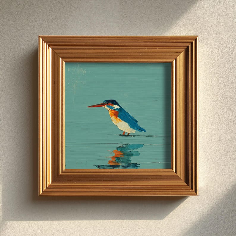 May include: A framed painting of a kingfisher bird. The bird has blue, orange, and white feathers and a long red beak. The painting is set against a teal background, with the bird's reflection visible in the water. The frame is gold.