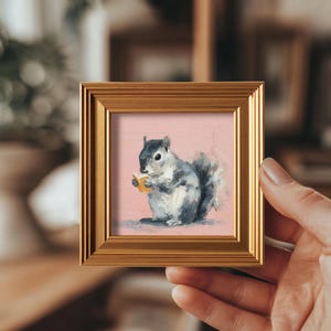 Peut inclure: Peinture encadrée d'un écureuil gris et blanc tenant un objet jaune, sur fond rose. L'œuvre est dans un cadre doré. La peinture présente un style texturé et au coup de pinceau.