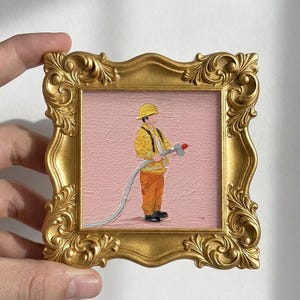 May include: A small, square painting in a gold ornate frame. The artwork depicts a firefighter in yellow gear holding a hose against a pink background. The frame is held by a hand.
