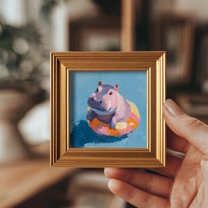 May include: A small, square painting in a gold frame depicts a purple hippo floating on a yellow and pink inflatable ring in blue water. The artwork is held by a hand, showcasing the framed art.