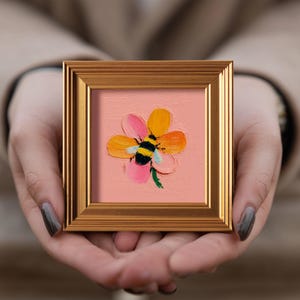 Könnte beinhalten: Ein kleiner, quadratischer, gerahmter Gemälde mit einer stilisierten Blume mit gelben, rosa und orangefarbenen Blütenblättern. Eine schwarz-gelbe Biene ist auf der Blume zentriert. Das Gemälde befindet sich in einem goldfarbenen Rahmen.